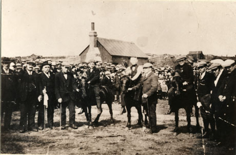 Photograph described as Easington Miners During Strike, Pony Racing at Welfare Park, showing three ponies with boys riding them and a group of men either side of them; a crowd of people can be seen immediately behind the ponies and men; behind the crowd, the roof and sides of a small house can be seen; and, in the distance, huts with the appearance of allotments