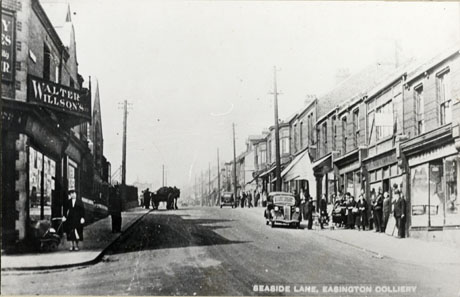 Postcard photograph entitled Seaside Lane, Easington Colliery looking up the road as the lane climbs the hill; facades of the shops can be seen on both sides of the road, including Walter Wilsons on the left-hand side of the road with the facade of a chapel beyond it; the names of the shops on the opposite side of the road cannot be determined; two motor cars and one cart can be seen in the road; a number of indistinct figures can be seen on the pavements, including a woman with a perambulator