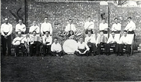 Photograph of nine men in shirt sleeves standing against a high brick wall; in front of them are nine men, also in shirt sleeves, sitting on deck chairs and holding musical instruments; the men are described as Easington Colliery Jazz Band, 1938
