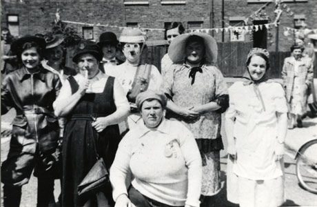 Photograph of a group of nine of people in fancy dress photographed in the road, with terraced houses in the middle distance; the people are described as being an Easington Colliery Carnival Group called Up Canada 1940; a number of indistinct figures can be seen behind those in fancy dress