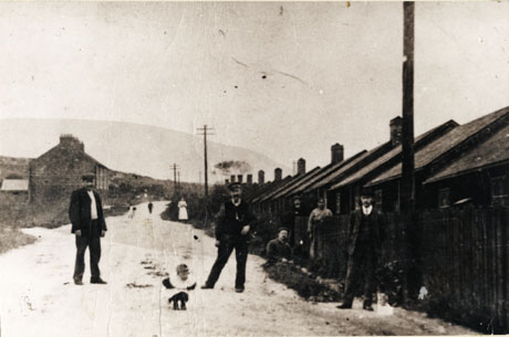 Photograph showing a row of low one-storied houses stretching away into the distance on the right- hand side of the photograph; the houses are behind fences; in front of the fences seven individuals may be seen indistinctly; on the opposite side of the road a short row of two-storied houses may be seen in the distance; the one-storied houses are described as Sinkers Huts, Easington Colliery(where Ascot Street now stands) 1910