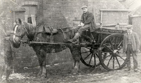 Photograph of a horse in the shafts of a cart with a man sitting on the front of the cart with his feet on the shafts of the cart; the two- wheeled cart bears the legend T. Monk Contractor; a man is standing near the rear of the cart, and a boy is holding the horse's head; the cart is standing near the back wall of a house in which a window can be seen