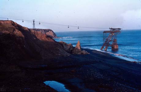 Photograph showing cliffs on the left of the photograph and sea on the right; containers can be seen on an aerial wire stretched between a pylon on the top of the cliffs and a structure in the sea; the photograph has been identified as being at Easington