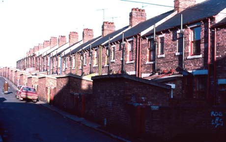 Photograph of the rear of a row of fifteen terraced houses running uphill away from the camera; two cars are parked facing away from the camera; the photograph has been identified as being in Easington colliery