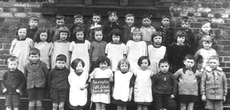 Photograph showing thirty children, aged approximately six years, posed against a brick wall; a child on the front row is holding a notice reading: Easington Church of England School Infants B 1927