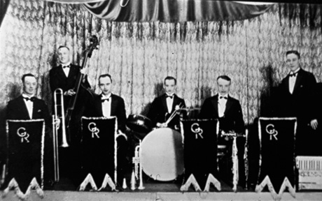 Photograph showing six men sitting and standing on a stage with stands with GR on them in front of them; the men are playing musical instruments and wearing dinner jackets; they have a curtain behind them; they have been identified as the Gilbert Ridley band