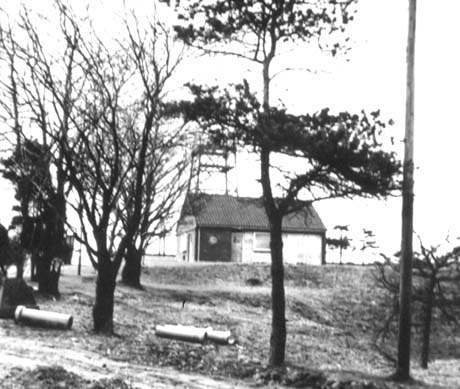 Photograph showing a small building on the brow of a hill with an indistinct pylon-like structure behind it; in the foreground are seven trees and pipes lying on the ground between the trees; the photograph has been identified as Pumping Station, Easington