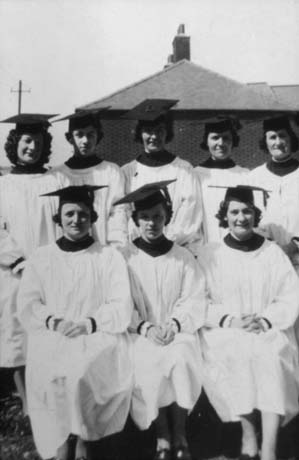 Photograph of eight people wearing mortar boards and surplices posed in two rows with the roof of a house behind them; they have been identified as Easington Colliery Choir