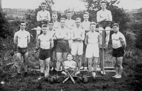 Photograph of eleven men in shorts and plimsolls posed in front of gymnastic bars with a view of countryside behind them; a boy aged approximately twelve years is sitting cross legged in front of the bars with two Indian clubs, two pairs of boxing gloves and a weight in front of him; they have been identified as members of the Health and Strength Club at Easington