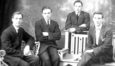 Photograph of four young men wearing suits and ties; two are seated at the left; one is seated on a bench over the back of which the fourth is leaning; the photograph has been taken in a photographer's studio; they have been identified as J. Barton; W. Roundling; F. Reynolds