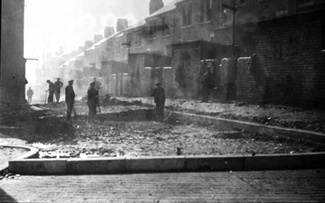 Photograph showing the rear of terraced houses on the right of the photograph and six men standing in the road laying the surface of the road; they are indistinct; at the front of the photograph a curb, defining the extent of the road, can be seen; the photograph has been identified as being in Easington Colliery
