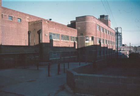 Photograph of the exterior of a large brick building of three storeys with the winding gear of the colliery behind it; a wall and a walkway can be seen in the foreground of the picture; the photograph has been identified as  Pit Baths, Easington Colliery