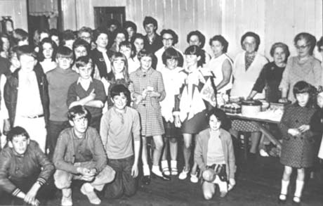 Photograph of a crowd of people and children standing in a hall with wooden walls; on the right of the picture a trestle table with cake tins on it and women standing behind it can be seen; the photograph has been identified as Church of The Ascension Fayre, Easington Colliery