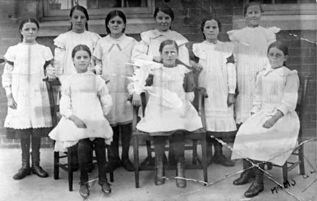 Photograph of six girls standing and three girls sitting on chairs in front of them; they are aged approximately twelve years and are wearing broderie anglaise pinafores, black stockings and boots; they have been described as being members of a cookery class at Easington Colliery