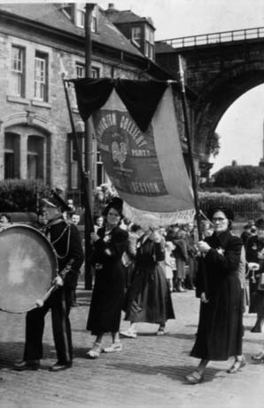 Photograph showing members of the Women's Section of the Easington Colliery Labour Party marching in Durham City at the bottom of North Road with the railway viaduct in the background; two women can be seen carrying the banner of the Women's Section with a man beating a drum preceding them; other members can be seen in the background