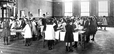 Photograph showing girls, aged approximately eleven years, standing at tables arranged in a large room, presumably a school hall; the girls are wearing gym slips and pinafores; there are lengths of cloth on the tables; two women can be seen at the left of the picture; the photograph has been identified as a Sewing Class in Easington Colliery