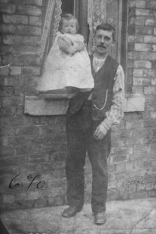 Photograph showing a man dressed in trousers, shirt and waistcoat, standing outside the window of a house holding a small child, aged approximately a year old, on his right arm; they have been identified as being in Easington Colliery