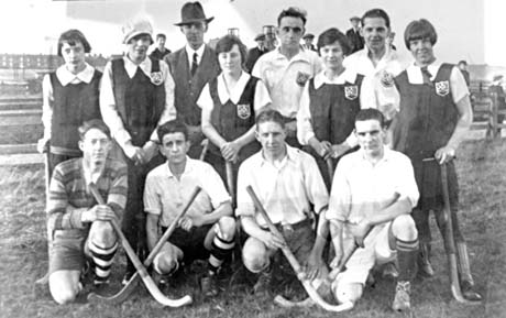 Photograph of five women in gym slips with badges on them and four men in shorts and boots; all eight are holding hockey sticks; a man in a suit and hat, and two men in shirts with the same badge on them, are also in the group; behind the group, six spectators and the winding gear of the colliery can be seen; they have been identified as a hockey team in Easington Colliery