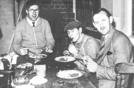 Photograph of three men sitting at a table with plates of food in front of them; one man is wearing a cardigan and tie and is sitting with his back to a rough brick wall at the head of the table; next to him is a young man in overalls and cap and next to him a man in a suit and tie; there is no cloth on the table, which also has an ashtray, a round tin and a tin of Carnation Milk on it; they have been identified as having Christmas Dinner in the Council Workshop; one of the men has been identified as Cliff Heslop