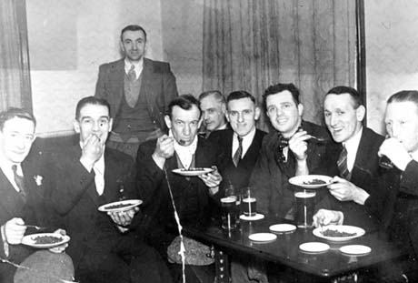 Photograph showing eight men sitting on benches round the wall of a club or public house with plates of peas in their hands; another man is standing behind them; a table with three glasses of beer on it is on the right of the photograph; the photograph has been identified as Carling Sunday at Easington Central Club, Easington Colliery