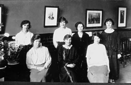 Photograph of seven women posed in a room with tiles half way up the wall and three pictures in frames behind them; they are wearing blouses and skirts and dresses; they have been described as Easington Colliery Staff