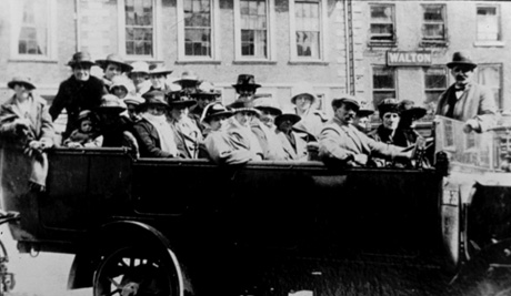 Photograph of approximately twenty three people in a motor charabanc, with buildings in the background, one of which has the word Walton on it; they are sitting in four rows looking at the camera; they have been identified as being on an outing from Easington Colliery