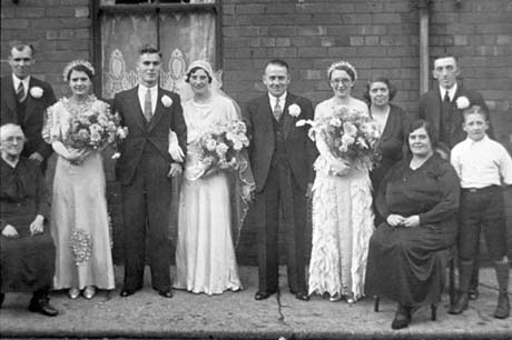 Photograph of a newly-married couple standing with two bridesmaids and four men in suits with flowers pinned to their coats; they are accompanied by three elderly women and a boy aged approximately twelve years; they are all posed in front of the window of a house; they have been identified as being in Easington Colliery