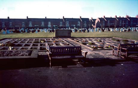 Photograph showing the memorial to the miners killed in the disaster in Easington Colliery in 1950; the memorial is a commemorative stone with eighty one spaces in front of it representing the number of miners who died; behind the memorial are graves in the cemetery and a line of terraced houses