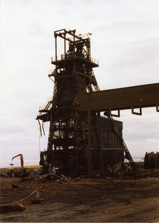 Photograph showing the winding gear of a colliery outlined against the sea; in the foreground is open cleared ground so it would appear that the buildings of the colliery are in the process of being demolished; the colliery has been identified as Easington Colliery