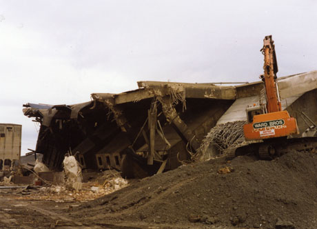 Photograph showing, at the right, a pile of soil with an earth moving vehicle on top with the words Instrip Demolition on it; in the centre of the photograph the remains of a large concrete structure, and, on the left, a large concrete building can be seen; the photograph has been identified as Easington Colliery