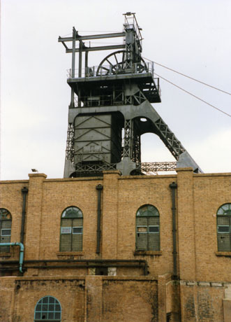Photograph of the exterior of a large brick building with round-topped windows; looming behind the building is the top part of the winding gear of a colliery; the colliery has been identified as Easington Colliery