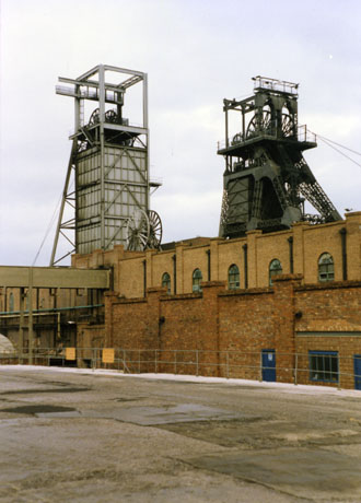 Photograph showing the exterior of the same brick building as in 185, taken from a longer distance away and from the right; the facade of the building can be seen and the top of two sets of winding gear; an expanse of concrete surface can be seen in the foreground along with a steel fence round the building on which notices concerning the demolition can be seen; the colliery has been identified as Easington Colliery