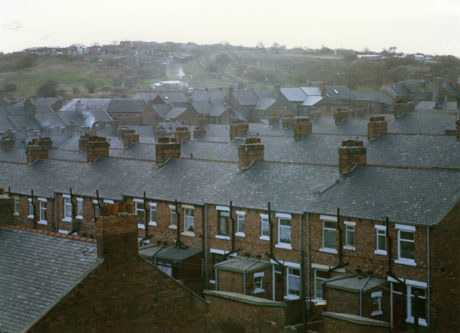 Photograph, taken from above, of the backs of a row of terraced houses; the roofs of further rows of terraced houses can be seen; beyond the roofs a hillside with, possibly, allotments on it can be seen; the place has been identified as Easington Colliery