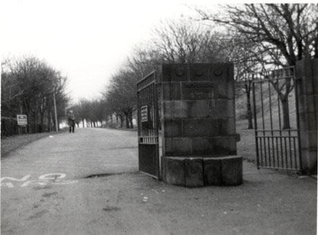 Photograph showing a stone pillar with two open gates of steel railings on either side; beyond the gates, a wide path of tarmac runs away from the camera between lines of trees on either of its sides; an indistinct man can be seen walking towards the camera down the path; the scene has been identified as being in Easington Colliery