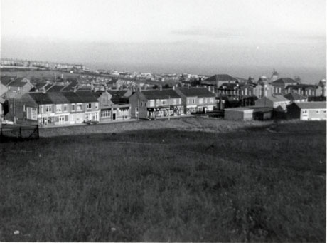 Photograph, taken from a hill, showing a parade of shops at the front of the settlement and the roofs of other buildings behind them; the details of the shops cannot be discerned; on the right of the picture, the Easington Junior School can be seen and beyond that the sea; the place has been identified as Easington Colliery