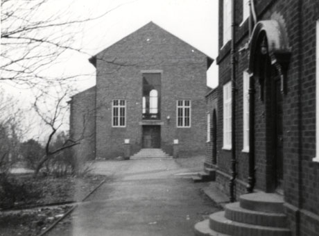 Photograph showing the side of a brick building with a long window in the centre of the building with a doorway and flight of steps leading to the doorway immediately below it; there is a window on either side of the central window; the side of the facade of another building with a canopy over the doorway and three round steps up to the doorway can be seen; the buildings have been identified as being in Easington Colliery