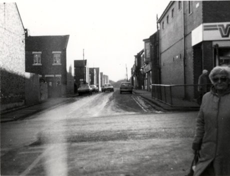 Photograph of a street running away from the camera; the ends of terraces of houses can be seen on the left where other streets abut the original street; on the right it is possible to see that there are shops on the street, but the details of these cannot be seen; three cars are parked and one is driving towards the camera; part of the figure of an elderly woman can be seen at the right of the photograph, which has been identified as being in Easington Colliery