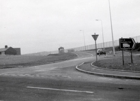 Photograph showing a road with grass on either side curving away to the right towards a wire fence, beyond which nothing can be seen; the end of a small building can be seen on the left and a sign reading N.C.B. Easington Colliery can be seen on the right; the photograph has been described as being in Easington colliery