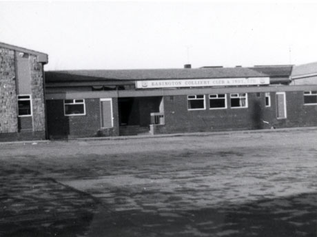 Photograph showing, across a concrete surface, the facade of a low building with a higher gable at one end; the building is of brick and the gable has stone facing; it bears a sign reading: Easington Colliery Club and Institute