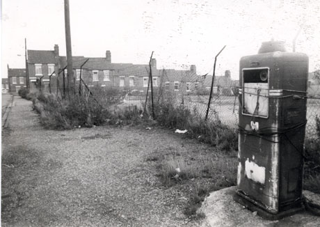 Photograph showing a disused and dilapidated petrol pump standing near a dilapidated fence of wire mesh with weeds growing at its base; beyond the fence is a terrace of houses; the photograph has been identified as Easington Colliery
