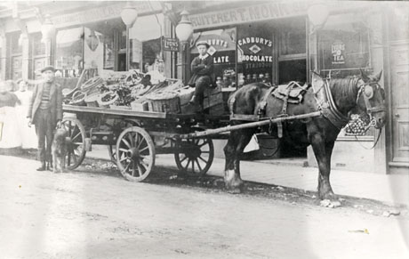 Photograph showing a horse between the shafts of a cart standing outside a shop, with the words Fruiterer T. Monk above the windows; one window has advertisements for Cadbury's Chocolate and for Lyons Tea on it and contains canisters and jars; the other window contains piles of fruit; the cart pulled by the horse contains boxes and baskets of fruit; a boy is sitting on a box on the cart and a man with a dog is standing by the cart at its back; three gas lamps can be seen between Monk's shop and that adjoining it; the shop adjoining has Tobacconist F. Douglas General Dealer written above it; the shops have been identified as being in Easington Colliery