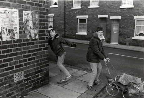 Photograph showing two boys, aged approximately thirteen years, dressed in anoraks and jeans, standing at the corner of a road; one is leaning on the wall and raising his foot, and the other is holding a string attached to a cart made of pram wheels; behind the boys, the facades of terraced houses across a road can be seen; the photograph has been identified as being in Easington Colliery