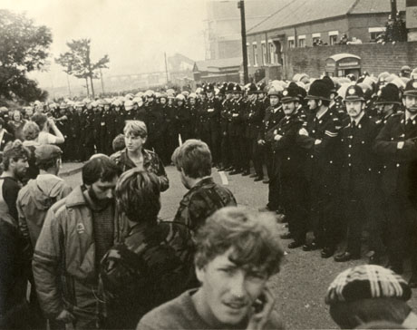 Photograph showing, on the right, a line of policemen, two or three deep, standing in front of the buildings of a colliery; on the left, is a group of men dressed in casual clothes, including camouflage jackets; behind the policemen, more policemen can be seen standing behind the walls of a building; it is possible that the photograph depicts scenes during the strike of 1984 in Easington Colliery