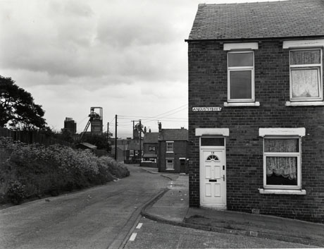 Photograph showing, on the right, the front of a terraced house with a road sign Angus Street on its wall; beyond the house, a road leads away from the camera with bushes on its left and the ends of three other streets on its right; at the end of the road, the winding gear and buildings of the colliery can be seen; the photograph has ben identified as Easington Colliery