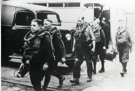 Photograph of five men in mines rescue gear walking in front of a motor vehicle; the two men in the rear are carrying a round piece of equipment with a narrow opening; behind the men, a man in uniform is standing near the open door of a van; the photograph has been identified as Easington Colliery