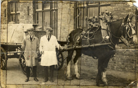 Photograph showing two men in suits, ties, caps, and overalls, standing near a horse which is between the shafts of a cart on which the words Durham Co-operative Bakery can be seen; the horse has decorations on its harness, as if it were taking part in a carnival; they all are standing in front of a large building with large windows; the photograph has been described as Easington Colliery