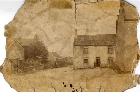 Photograph showing the exterior of a cottage with a tile roof, four windows and a doorway in the middle of the building; three indistinct figures are standing in the doorway; to the right in front of the cottage is the end of a house; to the left a garden wall, bushes and the roof and one wall of another house can be seen; the picture has ben identified as Easington Colliery