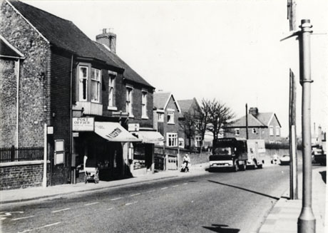 Photograph taken from the opposite side of the road of the exterior of a Post Office and cafe; and two shops with awnings; there are houses in the distance on the same side of the road as the shops; lorries can be seen in the road; the photograph has been identified as depicting the post office and a cafe in Easington Village Photograph, taken from the opposite side of the road, of the exterior of a Post Office and cafe; a name, possibly Alerton, can be seen on the awning of the building; another shop, also with an awning, can be seen next to the Post Office; detached houses can be seen in the distance further down the road on the same side; a motor lorry can be seen in the road and an indistinct figure with a push- chair can be seen on the pavement