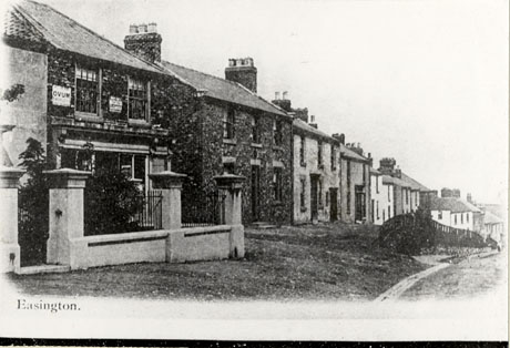 Photograph showing a row of houses of two storeys with plain facades running downhill away from the camera; the building closest to the camera is a shop, but it is not possible to see its windows; an advertisement for Ovum is on the wall; the scene has been identified as Rosemary lane in Easington village