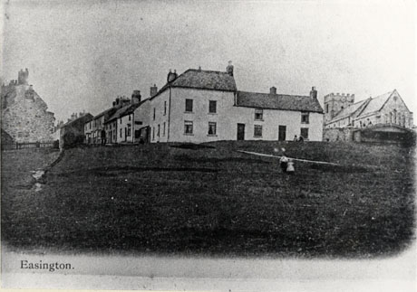 Photograph looking uphill towards the east end of the church of St. Mary, Easington, and the facade of a large house facing the Green, which occupies the foreground of the picture; a row of houses can be seen running at right angles to the end of the large house; small indistinct figures can be seen on the Green